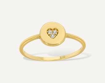 Könnte beinhalten: Ein goldener Ring mit einem kreisförmigen Design, in dessen Mitte sich ein herzförmiger Ausschnitt befindet. Das Herz ist mit drei kleinen, klaren Edelsteinen besetzt. Der Ring hat einen schmalen Band und eine polierte Oberfläche.