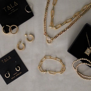 Peut inclure: Une collection de bijoux en or pr&eacute;sentant une vari&eacute;t&eacute; de styles, notamment des colliers, des bracelets, des boucles d'oreilles et des bagues. Les bijoux sont pr&eacute;sent&eacute;s sur une surface blanche avec des bo&icirc;tes noires.