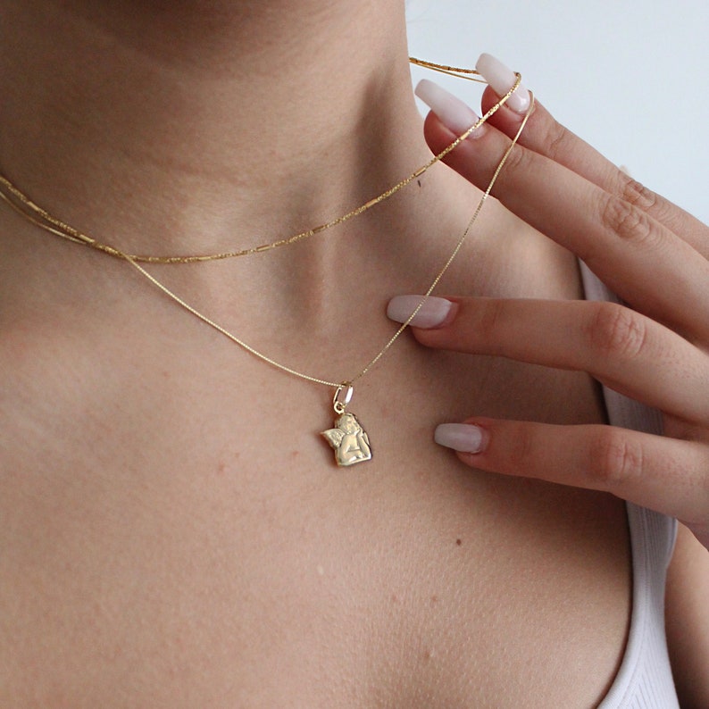 May include: A gold chain necklace with a small angel pendant. The necklace is layered with a second, thinner chain.