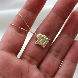 May include: A gold chain necklace with a small angel pendant. The angel is depicted with wings and a cherubic face.