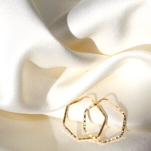 May include: A pair of gold-toned hexagonal hoop earrings. The earrings are on a white fabric background.