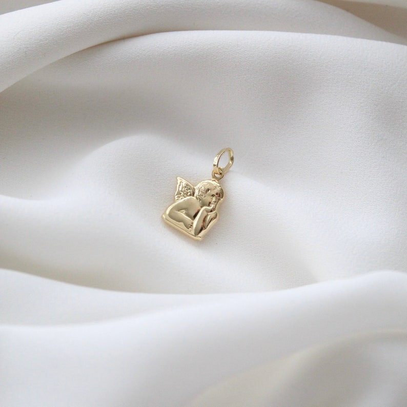 May include: A gold-toned pendant charm featuring a cherub angel with wings, holding its chin in thought.
