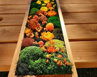 May include: A rectangular wooden planter box filled with a vibrant arrangement of preserved moss and dried flowers. The flowers are in shades of yellow, orange, and red, contrasting with the green moss. The box sits on a wooden surface.