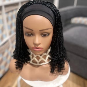 May include: A mannequin head displaying a black braided wig with a black headband. The wig features small, tight braids and curly ends. The mannequin is wearing a cream-colored, open-weave necklace.