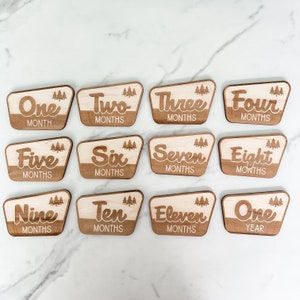 May include: Wooden milestone markers for a baby's first year. Each marker features a number from one to twelve, with the words "month" or "months" and a small tree graphic. The final marker says "One Year".