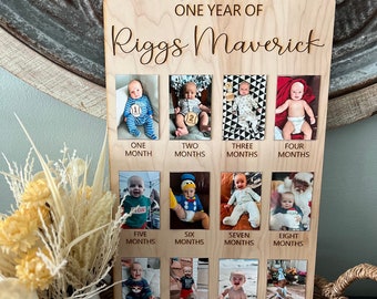 May include: A wooden photo display board with the text "ONE YEAR OF Riggs Maverick." It features twelve photo slots, each labeled with a month. The photos show a baby at different stages, from one to twelve months old. The board is made of light-coloured wood.