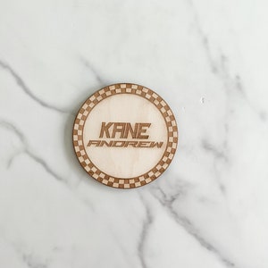 Puede incluir: Un posavasos redondo de madera con un borde a cuadros. El posavasos tiene el nombre "KANE ANDREW" grabado en el centro.