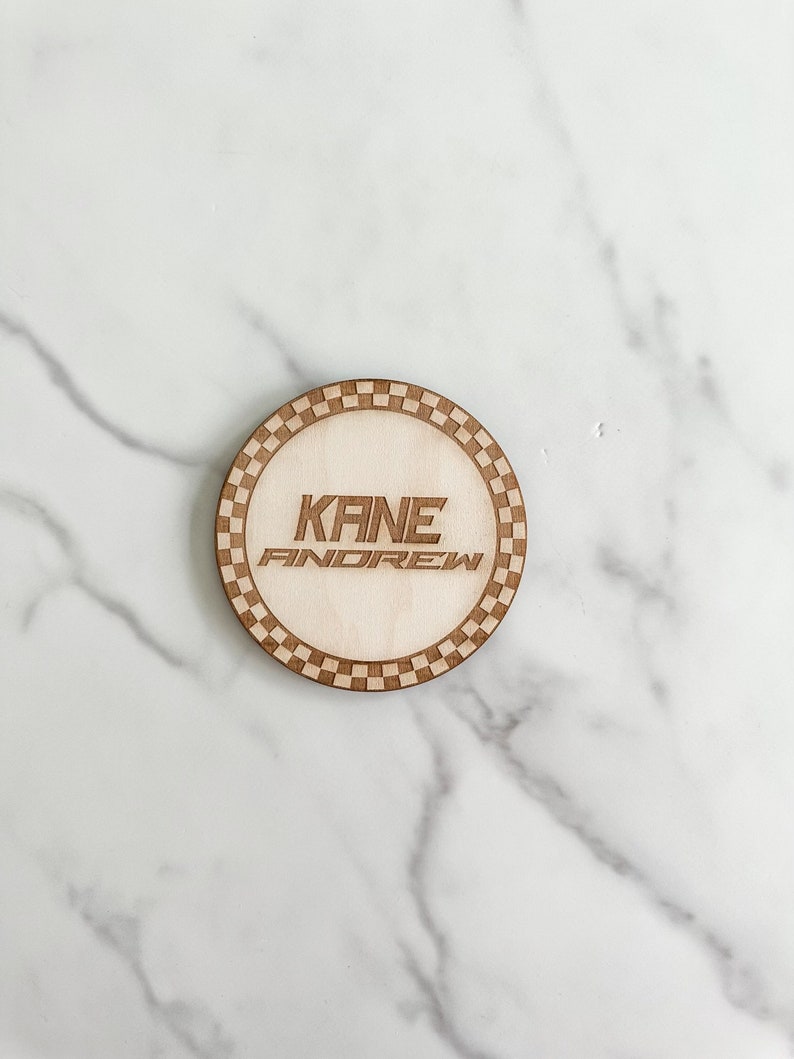 Puede incluir: Un posavasos redondo de madera con un borde a cuadros. El posavasos tiene el nombre "KANE ANDREW" grabado en el centro.