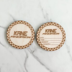 Puede incluir: Dos etiquetas redondas de madera para el anuncio de un beb&eacute; con un borde a cuadros. Una etiqueta dice "KANE ANDREW" y la otra etiqueta tiene espacios para rellenar la fecha, la hora, el peso y la longitud del beb&eacute;.