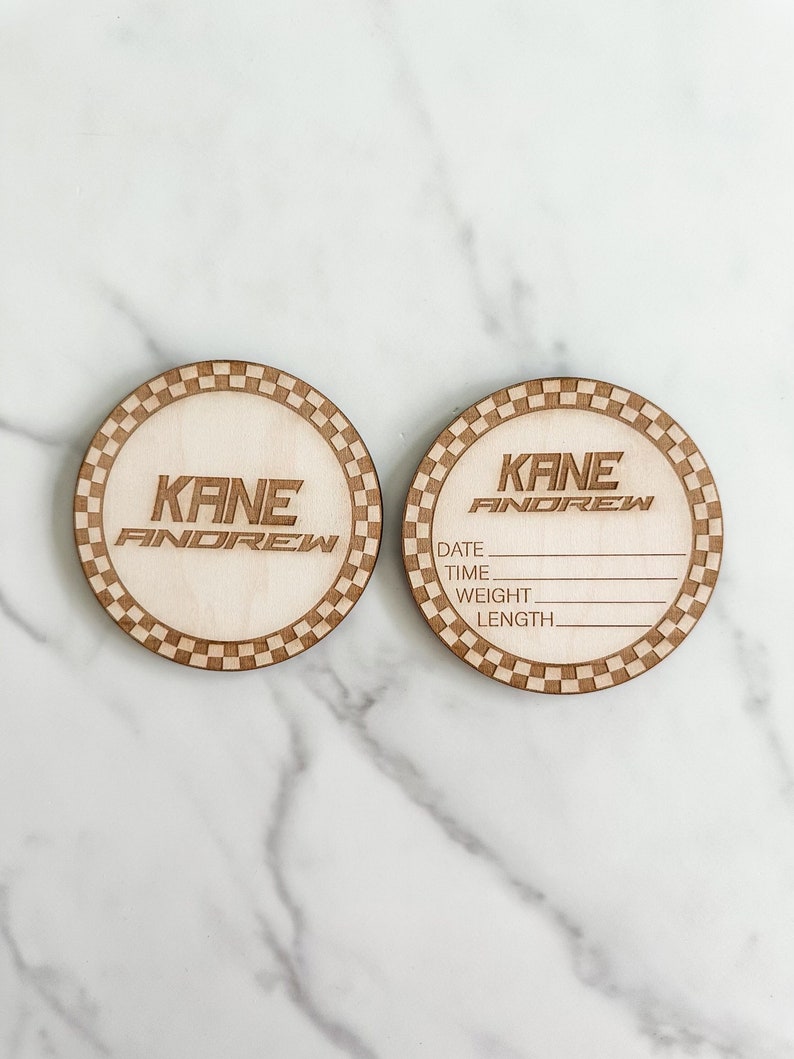 Puede incluir: Dos etiquetas redondas de madera para el anuncio de un beb&eacute; con un borde a cuadros. Una etiqueta dice "KANE ANDREW" y la otra etiqueta tiene espacios para rellenar la fecha, la hora, el peso y la longitud del beb&eacute;.