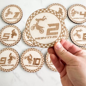 Puede incluir: Conjunto de diez discos de madera para hitos de beb&eacute; con tem&aacute;tica de motocross. Cada disco tiene un borde a cuadros blanco y negro y un n&uacute;mero del 1 al 10 con la palabra "MESES" debajo. Los discos presentan una silueta de un piloto de motocross realizando diversas acrobacias.