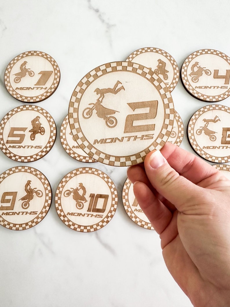 Puede incluir: Conjunto de diez discos de madera para hitos de beb&eacute; con tem&aacute;tica de motocross. Cada disco tiene un borde a cuadros blanco y negro y un n&uacute;mero del 1 al 10 con la palabra "MESES" debajo. Los discos presentan una silueta de un piloto de motocross realizando diversas acrobacias.