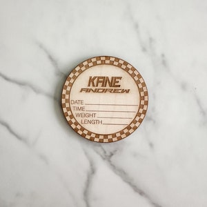 Puede incluir: Una placa redonda de madera con un borde a cuadros. La placa tiene el nombre "KANE ANDREW" grabado en ella. Tambi&eacute;n tiene las palabras "DATE", "TIME", "WEIGHT" y "LENGTH" grabadas en ella.