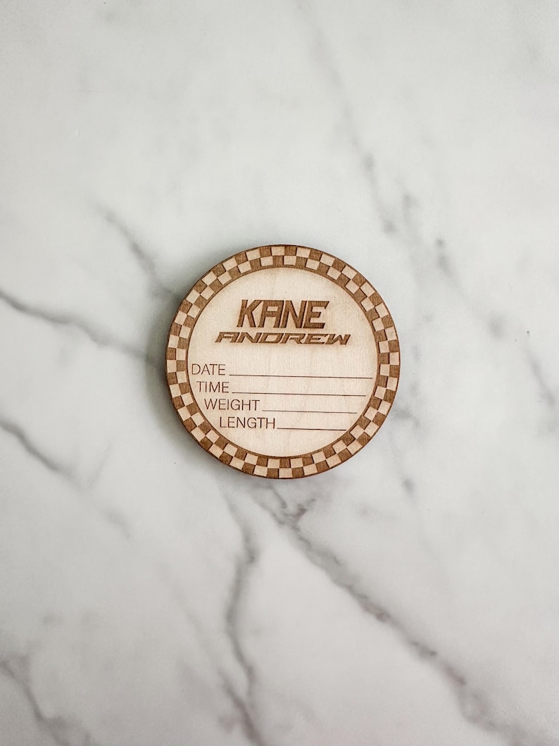 Puede incluir: Una placa redonda de madera con un borde a cuadros. La placa tiene el nombre "KANE ANDREW" grabado en ella. Tambi&eacute;n tiene las palabras "DATE", "TIME", "WEIGHT" y "LENGTH" grabadas en ella.