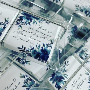 May include: A stack of small, clear boxes with white cards inside. The cards have a blue watercolor floral design and text that reads "Thank you for sharing our special day with us. Please keep us in your duas." and "Welcome to the Mikkah of..."