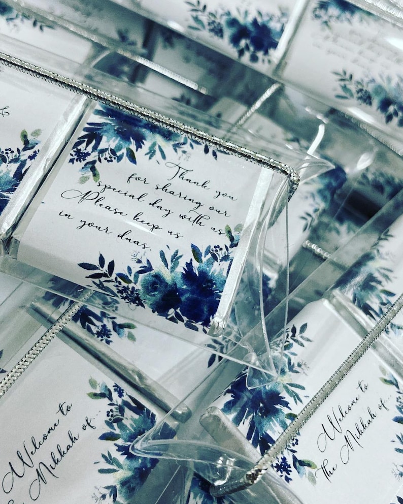 May include: A stack of small, clear boxes with white cards inside. The cards have a blue watercolor floral design and text that reads "Thank you for sharing our special day with us. Please keep us in your duas." and "Welcome to the Mikkah of..."