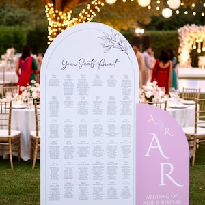 May include: A white wedding seating plan with a pink section. The sign says "Your Seats Await" and lists names and table assignments. The bottom right corner has the initials "A R" and the wedding details.