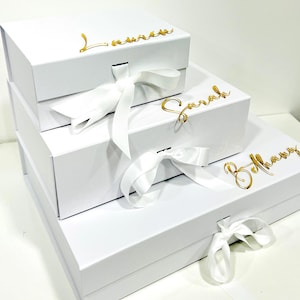 May include: Three white gift boxes with gold lettering and white satin ribbon bows. The boxes are stacked on top of each other. The top box says "Lauren", the middle box says "Sarah", and the bottom box says "Bethany".