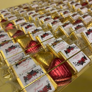 May include: A close-up of a box of small, individually wrapped chocolate hearts. The chocolates are gold and red and are decorated with floral designs. The box is labeled with the text "Nikkah of Arooj & Janeed" and "And we created you in pairs 78:8".