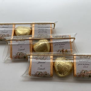May include: Three clear plastic favor boxes with gold trim and a gold heart-shaped chocolate inside each. The boxes have a white background with floral designs and text that reads "Junaid & Zainab" and "And we created you in pare 786".