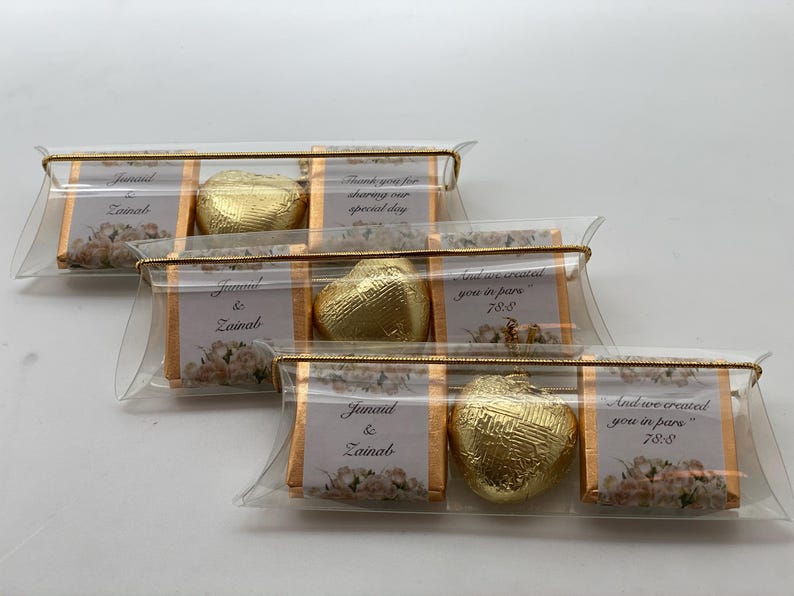 May include: Three clear plastic favor boxes with gold trim and a gold heart-shaped chocolate inside each. The boxes have a white background with floral designs and text that reads "Junaid & Zainab" and "And we created you in pare 786".
