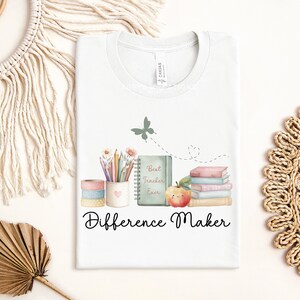 May include: White t-shirt with a watercolor design featuring pencils, books, an apple, and a butterfly. The text "Best Teacher Ever" is on a notebook, and "Difference Maker" is written below the design.