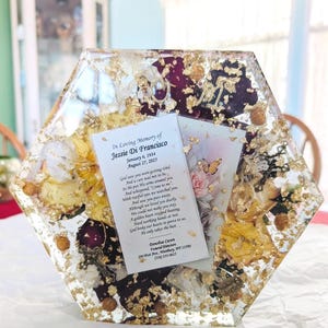Peut inclure: Une plaque commémorative hexagonale en résine contenant des fleurs séchées jaunes et rouge foncé, des flocons de feuilles d'or et une petite croix en métal. Une carte blanche est insérée au centre avec le texte : In Loving Memory of Jessie Di Francisco.