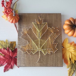 May include: A wooden sign with a string art design of a yellow and orange maple leaf. The leaf is made with copper wire and nails. The sign is surrounded by fall-themed decorations, including small pumpkins and fall leaves.