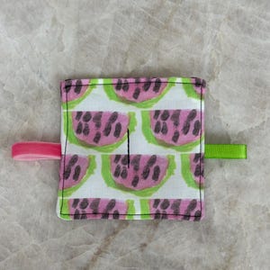 Puede incluir: Artículo de tela cuadrado con un diseño de sandía en rosa, verde y negro sobre un fondo blanco. Tiene un lazo elástico rosa en un lado y un lazo de cinta verde en el otro. El artículo es probablemente un pequeño accesorio o una pieza decorativa.