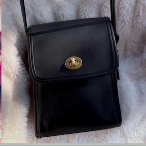 May include: A black leather crossbody bag with a rectangular silhouette and a fold-over flap. The bag features a gold-toned metal turn-lock closure centered on the flap and a thin black leather shoulder strap attached to the sides.