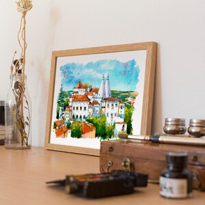 May include: A framed watercolor painting of a white building with red tile roofs and blue sky. The building is surrounded by trees and a green field. The painting is in a wooden frame.
