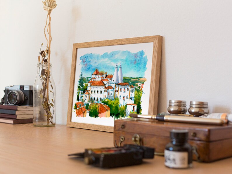 May include: A framed watercolor painting of a white building with red tile roofs and blue sky. The building is surrounded by trees and a green field. The painting is in a wooden frame.