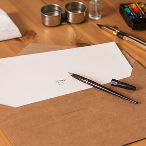 May include: A brown envelope with a white sheet of paper inside. A black fountain pen is lying on top of the envelope. The pen has a gold trim and a black cap. The paper has a handwritten signature on it.