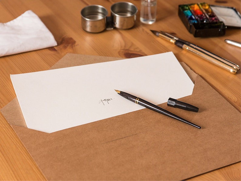 May include: A brown envelope with a white sheet of paper inside. A black fountain pen is lying on top of the envelope. The pen has a gold trim and a black cap. The paper has a handwritten signature on it.