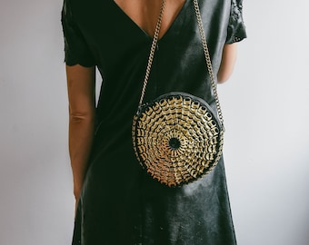 May include: A black circular handbag with a gold chain strap. The bag features a circular pattern of gold-coloured elements. The person is wearing a black dress.