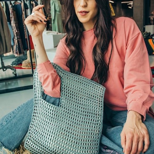 May include: A silver tote bag made from recycled aluminum can tabs. The bag has a rectangular shape with a handle and a perforated design. The person in the image is wearing a pink long-sleeve shirt and blue jeans.