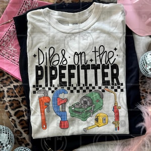 以下が含まれることがあります： 「Dibs on the Pipefitter」の文字と配管工の工具のイラストが描かれた白いTシャツ。工具には、レンチ、パイプカッター、パイプねじ切り機、巻尺、トーチが含まれます。Tシャツは黒いTシャツの上にあります。