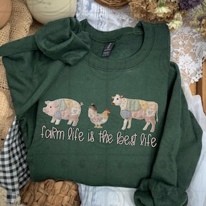 May include: Dark green sweatshirt with an embroidered design of a pig, chicken, and cow, each with patchwork patterns. The text "farm life is the best life" is embroidered below. The sweatshirt is folded.