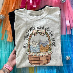 May include: Cream-colored t-shirt with a graphic of a basket filled with chickens. The basket has a floral bow and the text "Forget about candy, bring chickens instead." A black t-shirt is folded behind it.