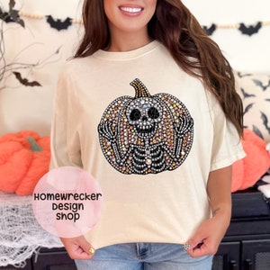 May include: A beige t-shirt with a rhinestone pumpkin design. The pumpkin is made of rhinestones in various shades of orange, clear, and black. The pumpkin has a skeleton inside with a smiling face.