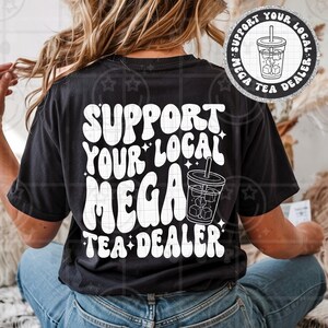 May include: A black t-shirt featuring white text that reads SUPPORT YOUR LOCAL MEGA TEA DEALER in a wavy font. The design includes a graphic of a plastic cup filled with iced tea and a straw, surrounded by small star accents on the back.