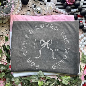 May include: A stack of three t-shirts in gray, pink, and white. The gray shirt features the text "For God So Loved The World" in a circular design, with a bow graphic and the text "John 3:16".