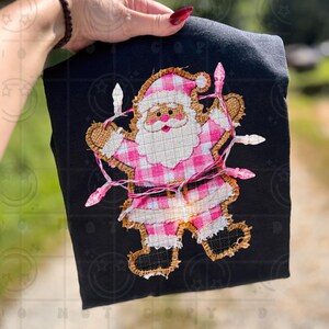May include: Black t-shirt featuring a Santa Claus design with a pink and white plaid pattern. The Santa Claus is outlined with burlap and adorned with pink string lights. A festive holiday-themed graphic.
