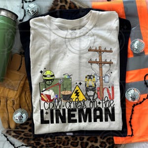 May include: Cream-colored t-shirt with the text "I call dibs on the LINEMAN" and illustrations of lineman tools and equipment. The design includes a utility pole, hard hat, and safety gear. The shirt is folded on top of a black shirt.