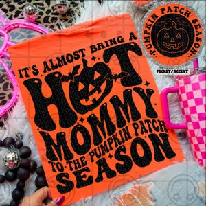 May include: Bright orange t-shirt with the text "IT'S ALMOST BRING A HOT MOMMY TO THE PUMPKIN PATCH SEASON" in black. A circular graphic with a jack-o'-lantern is on the upper right. A pink handled tumbler is in the background.