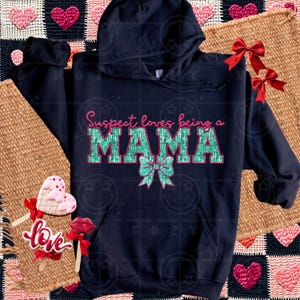 May include: A navy blue hoodie featuring the text "Suspect loves being a MAMA" in pink and green, with a bow graphic. The sweatshirt is presented on a textured surface, with decorative elements. The design is eye-catching.