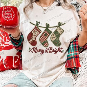 Puede incluir: Una camiseta color crema con un gráfico de tres calcetines navideños colgantes en tonos rojo, verde y cuadros. Debajo de los calcetines, el texto Merry & Bright está impreso en una fuente cursiva de color rojo oscuro. La camiseta se lleva con una camisa de franela a cuadros.