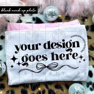 Puede incluir: Camiseta blanca con el texto "your design goes here" en negrita negra. El diseño incluye estrellas negras y lazos. La camiseta está doblada sobre una camiseta rosa. El fondo incluye bolas de discoteca y un estampado de piel sintética.