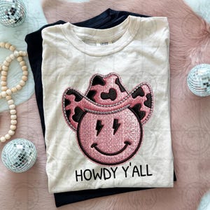 Puede incluir: Camiseta color crema con un gráfico de cara sonriente con sombrero de vaquero rosa y negro y el texto "HOWDY Y'ALL". La camiseta está superpuesta a una camiseta negra. Bolas de discoteca decorativas y cuentas de madera en el fondo.