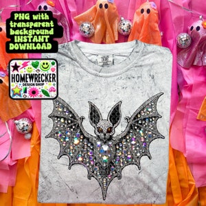 May include: A white t-shirt with a distressed look featuring a rhinestone bat design. The bat is black with red eyes and is surrounded by iridescent rhinestones. The t-shirt is on a pink and orange background.